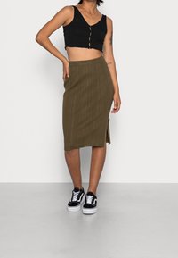 Černý cropped top s detaily knoflíků, doplněný o šedozelenou ribbed midi sukni s bočním rozparkem. Obuté jsou černobílé tenisky.