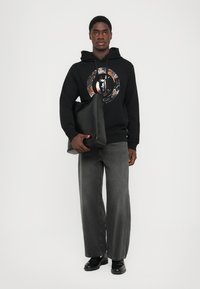 Hombre con sudadera negra con gráfico circular, pantalones anchos grises, zapatos negros, sosteniendo una gran bolsa tote negra, de pie contra un fondo blanco.