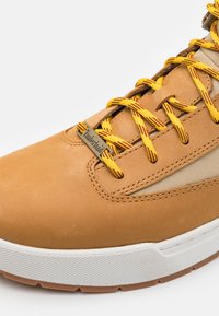 Bruna lädersneakers med gula och röda snören, nätaccenter och en vit gummisula med ett texturerat greppmönster.