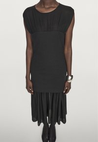 Schwarzes Kleid mit bodenlangem Oberteil und Falten, kurzen Ärmeln und einem ausgestellt, gestuften Rock. Weiche Textur, knielang.