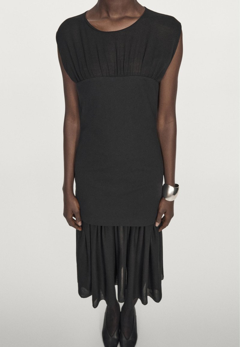 Schwarzes Kleid mit bodenlangem Oberteil und Falten, kurzen Ärmeln und einem ausgestellt, gestuften Rock. Weiche Textur, knielang.