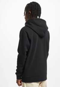 Persona con pelo trenzado negro, vistiendo una sudadera con capucha negra y pantalones beige, de espaldas frente a un fondo blanco.
