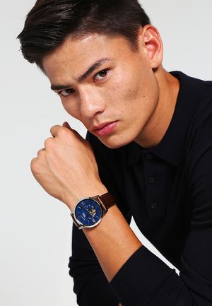 Montre avec bracelet en cuir brun, boîtier rond en acier inoxydable et cadran bleu mettant en valeur des accents argentés et un fond transparent.