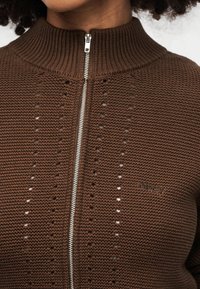 Καφέ πλεκτός πουλόβερ με ψηλό γιακά, ασημένιο φερμουάρ και λεπτομέρειες με υφαντό σχέδιο. Το λογότυπο της μάρκας «obey» εμφανίζεται διακριτικά στο εμπρός μέρος.