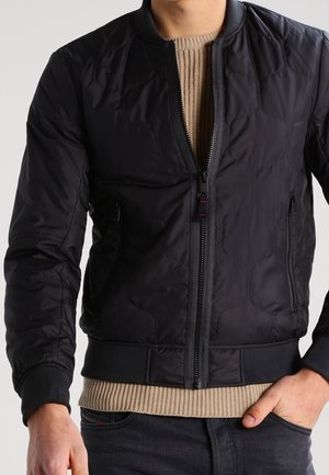 Man wearing a black quilted bomber jacket over a beige knit sweater, with one hand in dark jeans pocket.