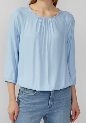 Blouse bleu clair à manches longues avec encolure froncée portée rentrée dans un jean bleu délavé moyen sur une personne debout devant un fond uni.