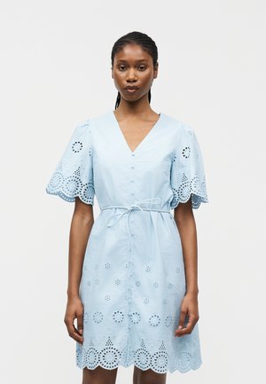 Kvinde iført en lyseblå kjole med hulmønstre, korte puffede ærmer, knappet forside og et bundet bælte i taljen mod en ensfarvet baggrund.