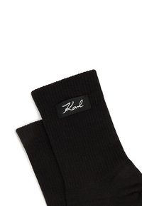 Czarne prążkowane skarpetki z prostokątną czarną metką z białym napisem "Karl" w stylu pisma odręcznego. Materiał wydaje się miękki i elastyczny.