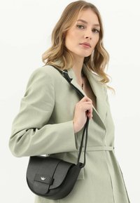 Czarna skórzana torebka na ramię o zakrzywionym kształcie, teksturowanej powierzchni i klapie z metalowym, markowym akcentem. Dołączony regulowany pasek.