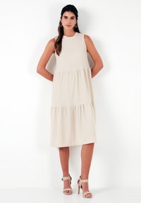 Beige sleeveless dress with a tiered design, made from lightweight fabric. Features a round neckline and falls to mid-calf length.