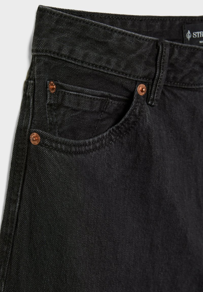 Jeans en denim noir avec une surface légèrement texturée, présentant des rivets de couleur cuivrée, une poche avant et un design de taille classique.