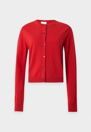 Frau mit langen dunklen Haaren trägt eine rote, geknöpfte Strickjacke, graue weite Jeans und schwarze spitze Schuhe, steht mit einer Hand in der Tasche.