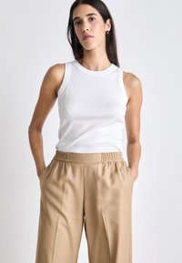 Λευκό ριμπ αμάνικο τοπ συνδυασμένο με μπεζ παντελόνια ευρείας γραμμής που διαθέτουν ελαστική μέση. Το μοντέλο στέκεται με τα χέρια στις τσέπες σε έναν απλό φόντο.