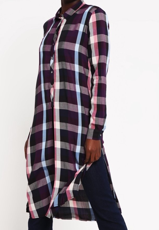 Personne portant une robe chemise à manches longues boutonnée, à carreaux avec des rayures roses, bleues, noires et blanches, associée à un pantalon foncé.