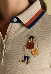 Beiger Poloshirt mit einem Kragen in Marineblau und Roststreifen, verziert mit einem gestickten Fuchs in rotem Pullover mit "J", cremefarbene Hose und dunkle Stiefel.