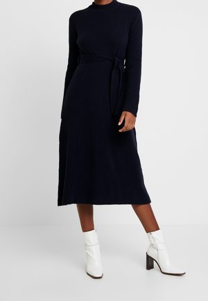 Vrouw draagt een navy blauwe gebreide jurk met lange mouwen, een taillekoord en witte enkellaarsjes, staand tegen een effen lichte achtergrond.