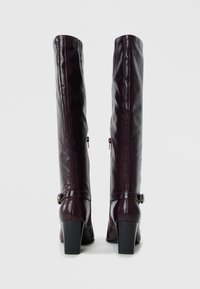 Knee-high burgundy leather boots with a glossy finish, square heels, and side zippers; adjustable straps with decorative buckles.