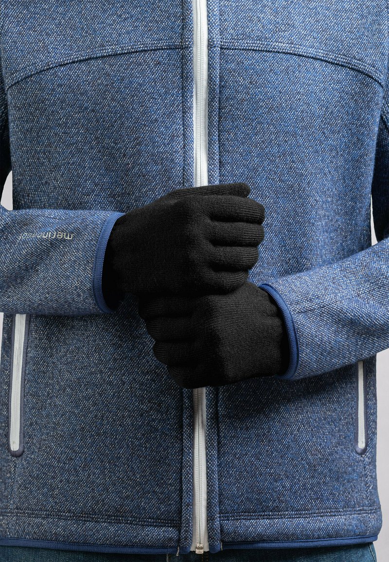 Des gants en tricot noirs sur une personne portant une veste zippée bleue texturée avec des accents blancs et une étiquette visible au niveau du poignet.