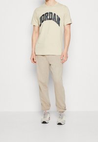Beżowa bawełniana koszulka z czarno-zielonym nadrukiem w kratkę "JORDAN", zestawiona z beżowymi spodniami dresowymi i szarymi trampkami z czerwonymi akcentami.