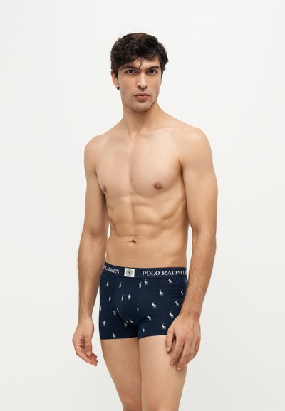 Νεαρός άνδρας που φοράει σκούρα μπλε boxer Polo Ralph Lauren με λευκά λογότυπα, στέκεται μπροστά σε απλό ανοιχτό φόντο.