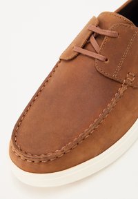 Hnedá kožená topánka s hladkým povrchom, s okrúhlou špičkou a jemným prešívaním. Obsahuje bielu gumovú podrážku a šnúrky.