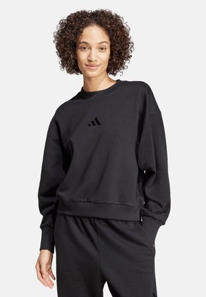 Donna con capelli ricci che indossa una felpa nera Adidas a manica lunga e pantaloni neri coordinati, in piedi con una mano in tasca.