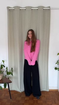 Rózsaszín kötött pulóver hosszú ujjakkal, széles fekete nadrággal párosítva. A háttérben világoszöld függöny és fa padló látható.