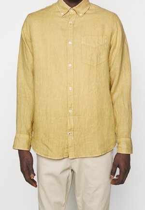 Homme portant une chemise jaune à manches longues avec une poche poitrine et un pantalon beige, debout devant un fond uni.