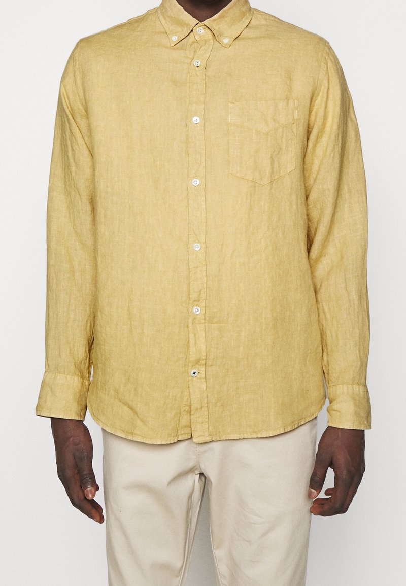 Homme portant une chemise jaune à manches longues avec une poche poitrine et un pantalon beige, debout devant un fond uni.