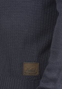 Maglione lavorato a maglia blu navy con un motivo testurizzato e una toppa in pelle etichettata "Solid" sul lato sinistro inferiore.