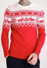 Maglione di Natale rosso e bianco con motivi di renne e fiocchi di neve indossato da una persona tatuata con le mani in tasca.