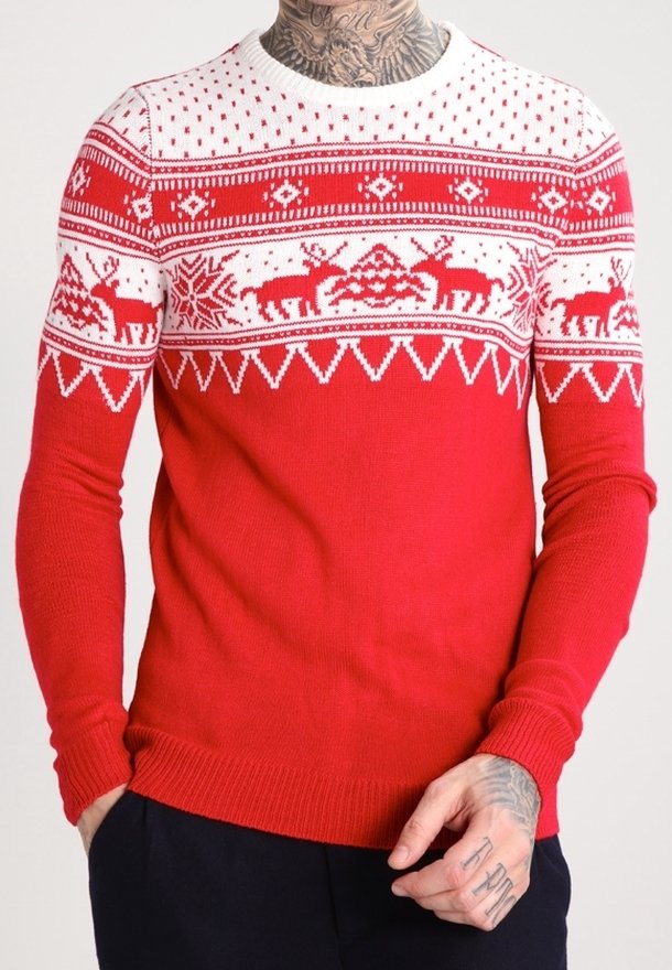 Maglione di Natale rosso e bianco con motivi di renne e fiocchi di neve indossato da una persona tatuata con le mani in tasca.