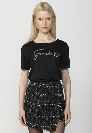 Camiseta negra de manga corta con un diseño de logo brillante. Combinada con una falda a cuadros en blanco y negro con textura, detalles de flecos y botones.