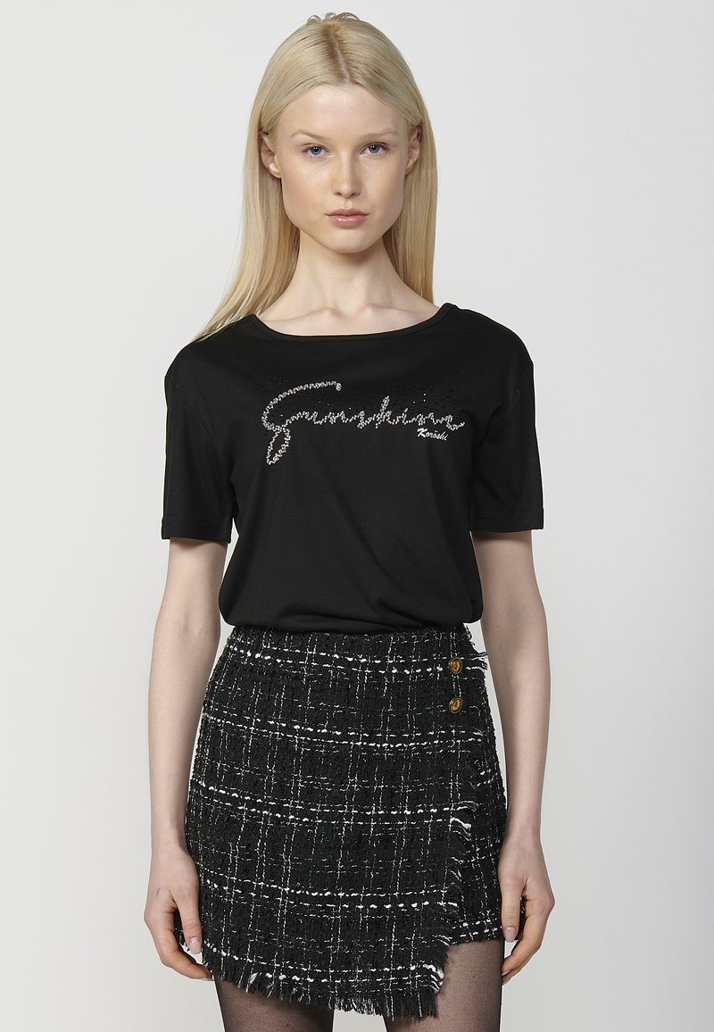 Camiseta negra de manga corta con un diseño de logo brillante. Combinada con una falda a cuadros en blanco y negro con textura, detalles de flecos y botones.