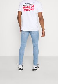 Camiseta blanca de manga corta con la frase "SUEÑOS HECHOS DE CROMO" impresa en azul y rojo, combinada con jeans pitillo azul claro y zapatillas negras y blancas.