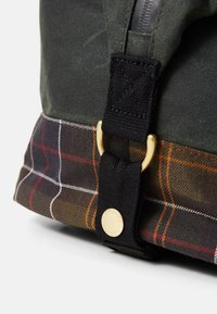Close-up of green fabric bag with brown plaid base, black strap, and gold metal ring with round gold button stamped "Barbour."