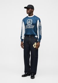 Μπλε και λευκό αθλητικό πουκάμισο με μακριά μανίκια, με εκτύπωση λογότυπου και κειμένου, συνδυασμένο με σκούρο τζιν παντελόνι που διαθέτει χρυσή κεντητή σχεδίαση.