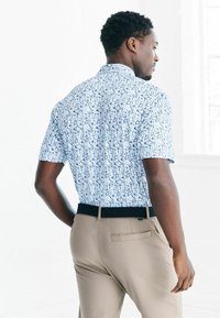 Kurzärmliges Hemd mit Knöpfen in Blau und Weiß mit einem geometrischen Muster, kombiniert mit beigen Hosen mit einem dunklen Taillenbund und einer Gesäßtasche.