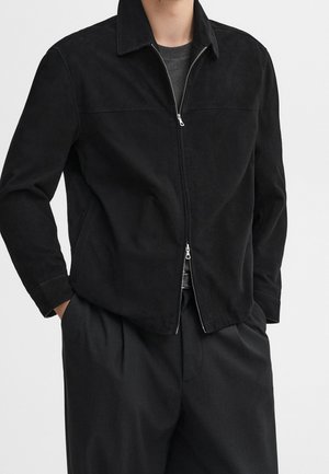 Homme portant une veste noire zippée par-dessus une chemise gris foncé, les mains dans les poches d'un pantalon noir ample.