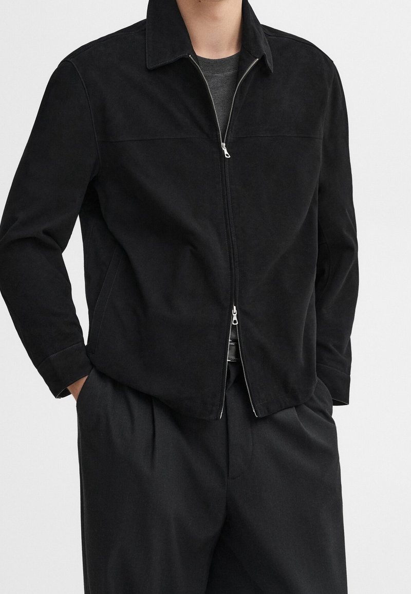 Homme portant une veste noire zippée par-dessus une chemise gris foncé, les mains dans les poches d'un pantalon noir ample.