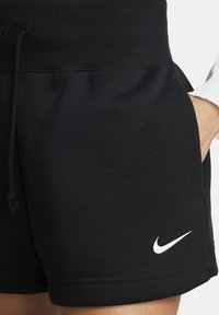Svarta sportshorts med ribbats midjeband och dragsko, med en vit Nike-swoosh-logotyp nära fållen, hand i sidfickan.