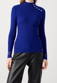 Femme portant un pull ajusté bleu côtelé avec des boutons argentés sur l'épaule et un pantalon en cuir noir, debout devant un fond uni.