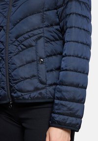 Veste matelassée en duvet bleu marine avec fermeture éclair, dotée d'une poche latérale à bouton-pression et de détails contrastants à l'ourlet.