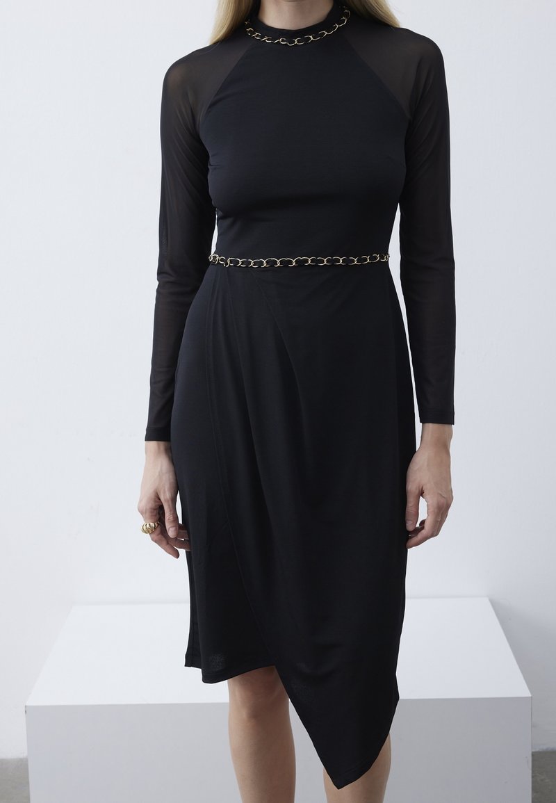 Femme portant une robe noire à manches longues avec un détail chaîne au col et à la taille, un ourlet asymétrique et une bague en or à son doigt.