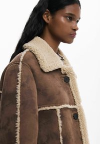 Giacca in shearling marrone con un colletto cremoso e accogliente, caratterizzata da grandi bottoni e cuciture a contrasto. Tessuto testurizzato con una forma ampia.