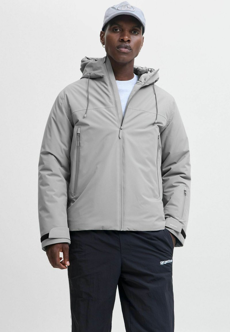 Grau gepolsterte Jacke mit Kapuze, zwei seitlichen Taschen und verstellbaren Bündchen. Kombiniert mit schwarzen Hosen, die ein Logo am Bein haben.