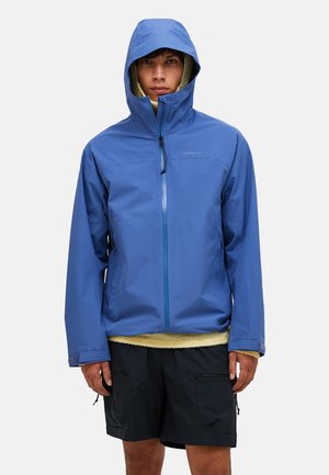 Ung mand iført en blå hætte-regnjakke over en beige sweater og sorte shorts, stående mod en enkel lys baggrund.