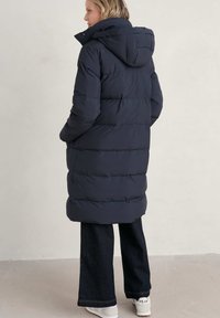 Abrigo tipo puffer en color navy con capucha, con un diseño de acolchado y bolsillos laterales. Se lleva sobre unos pantalones vaqueros de campana oscuros y zapatillas blancas.