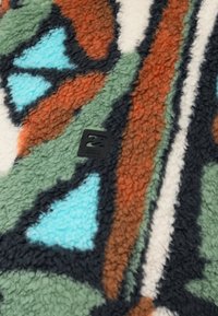 Viacfarebná fleecová prikrývka s abstraktnými tvarmi v zelenej, oranžovej, čiernej a modrej farbe. V jednom rohu je malá čierna štítková značka.