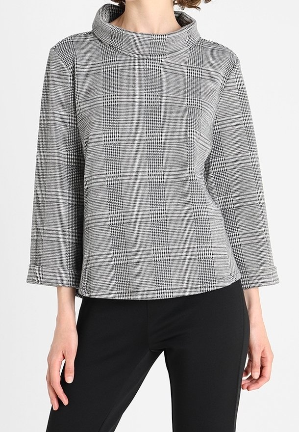 Femme portant un haut à manches longues à col montant gris à carreaux, associé à un pantalon noir ajusté, sur un fond uni.